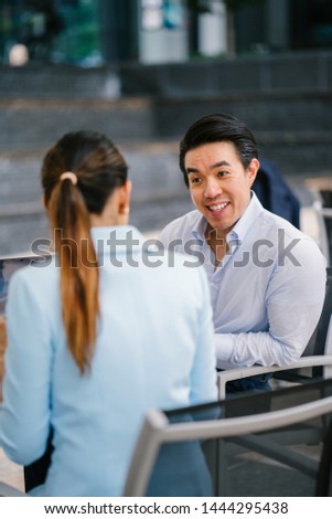A young, handsome and well-groomed Asian man in a shirt and pants is getting interviewed by a professional woman in a blue suit during the day. He is smiling and talking confidently. 