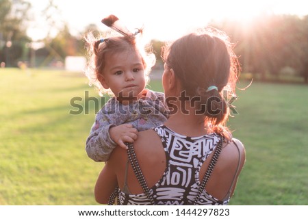 Young mother carrying kid in arms. Mom holding child outdoors in urban park in golden hour. Woman hugging toddler with feather in hair walking away from camera in nature at sunset. Royalty-Free Stock Photo #1444297823