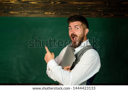 Education, school, people concept. Male teacher in classroom with laptop. Happy teacher at auditorium. Teacher in classroom near blackboard pointing at copy space. Teacher giving lesson to students.