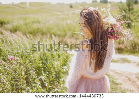 A beautiful young woman stands with her back in a frame in a pink dress looking away and smiling, holding a bouquet of wild flowers in her hands and smiling