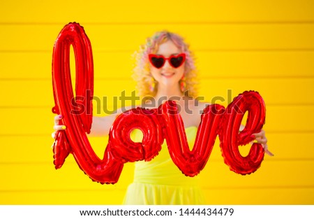A happy bright girl in sunglasses in the shape of hearts holds in her hands an inflatable ball with the word Love. The concept of celebrating Valentine's Day. Woman on a yellow background.