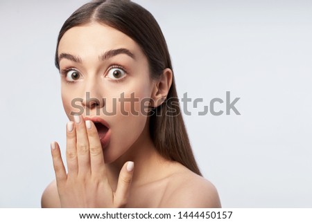 Waist up portrait of beautiful woman with perfect skin looking at camera and keeping hand while closing her mouth. Isolated on light gray background