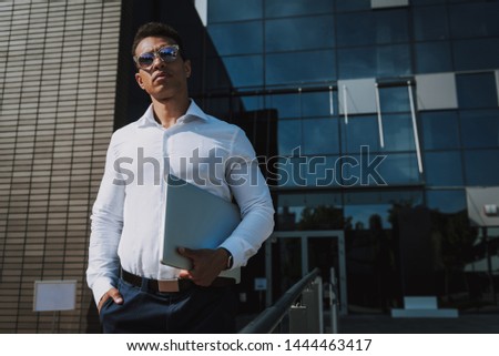Focus on elegant man in sunglasses standing near business center. He is carrying notebook and looking forward. Copy space in right side