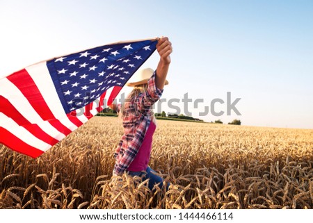 Concept of freedom and liberty. Attractive blonde female walking through wheat field and holding USA waving flag. Powerful economy and agriculture of the United States of America. Proud on her country