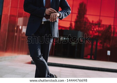 Focus on elegant man standing on front of business center. He is looking at smart watch and checking time. Copy space in right side