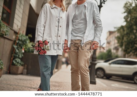 Cropped photo of romantic man and lady holding hands while walking in the city with flower