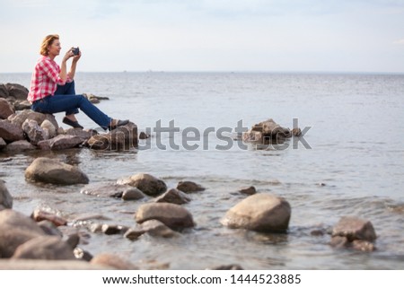 Caucasian woman sitting on stones near the water. Making pictures with cellphone, calm sea