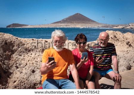 Three caucasian senior people sitting on a bench under the bright sun looking at their mobile phone.  Beautiful seascape behind them. Blue sky and sea with white waves. Paradise for surfers