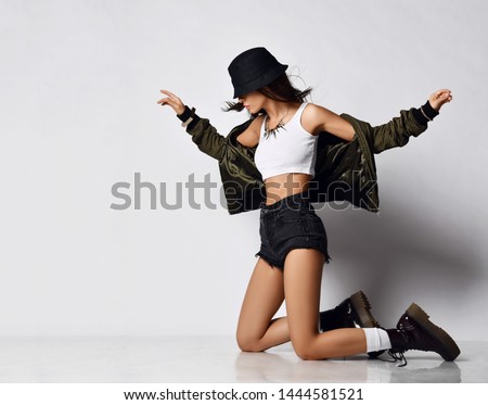 Young beautiful hipster girl sitting on her knees full body in shorts brown spring jacket, modern stylish boots and hat happy smiling on gray background