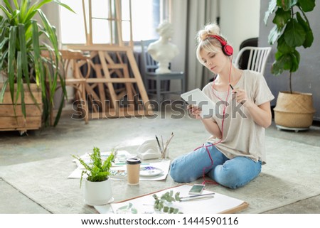Portrait of smart and creative woman looking at piece of pretty drawings with big concern and worry. Sad lady holding painting brush and sitting on floor of big studio. Arts and Crafts concept