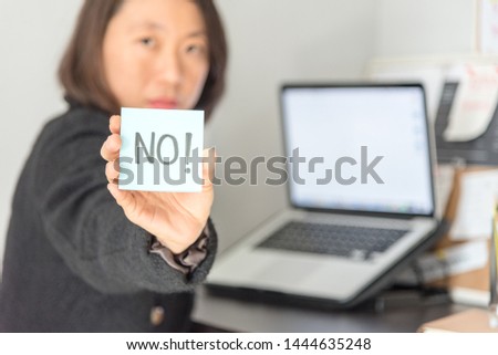 A woman sitting at a desk in working dress holding a paper note hands writing with the inscription No in front of her. No concept.