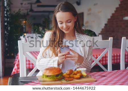 Girl with a long hair sitting in a restaurant. Girl food blogger takes a picture on the phone from social networks. Woman shooting food in a restaurant. Makes a photo of burger and fried potatoes