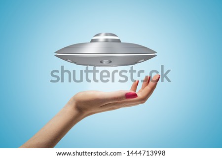 Side closeup of woman's hand and little UFO in air above her palm on light blue gradient background. Encounters with UFOs. Seeing is believing. Mysterious phenomena.