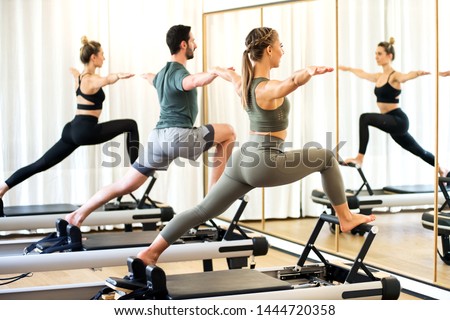 Class in a gym doing pilates standing lunges on reformer beds to stretch and tone the muscles reflected in a wall mirror Royalty-Free Stock Photo #1444720358