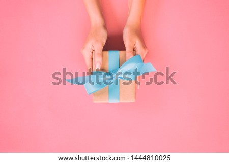 Woman hands give wrapped valentine or other holiday handmade present in paper with blue ribbon. Present box, decoration of gift on pink table, top view with copy space. Royalty-Free Stock Photo #1444810025