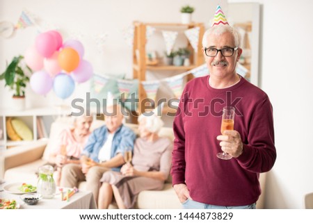 Casual aged man with flute of champagne on background of his friends on couch at birthday party
