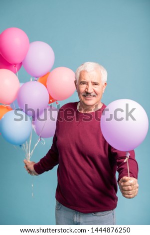 Cheerful mature man giving you one balloon from bunch while posing on blue background