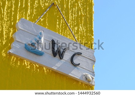 WC street sign. The sign of toilet on the white wooden board with small sail boat. WC outdoor sign hanging on a yellow scratched wall