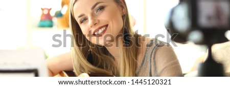 Happy Guitar Performer Recording Musical Vlog. Caucasian Cute Woman Sitting and Playing Music. Joyful Blogger Filming on Camera at Apartment. Cute Girl Bass Player Broadcast Rehearsal