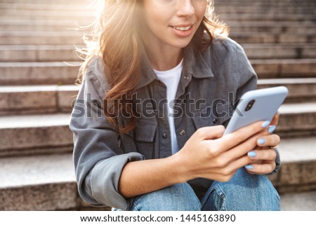 Cropped image of a cheerful young girl sitting on stairs outdoors, using mobile phone
