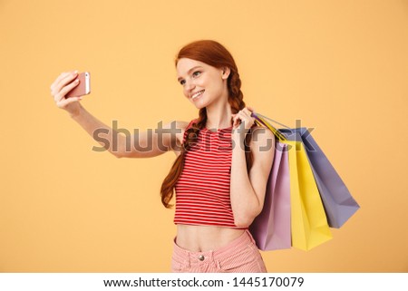 Image of a smiling young pretty redhead woman posing isolated over yellow background holding shopping bags take a selfie by mobile phone.