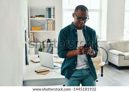 Using new mobile app. Handsome young African man in shirt using smart phone while standing in the office Royalty-Free Stock Photo #1445181932