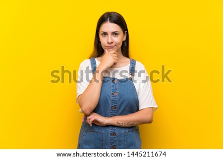 Young woman in dungarees over isolated yellow background thinking
