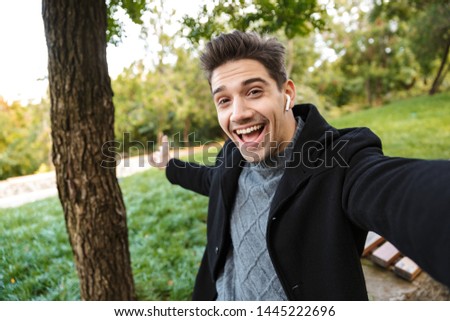 Image of optimistic pleased young man in casual clothing walking outdoors in green park take a selfie by camera listening music with earphones showing copyspace.