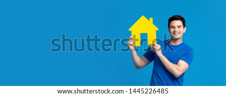 Smiling handsome caucasian man holding  house model isolated on blue banner background with copy space