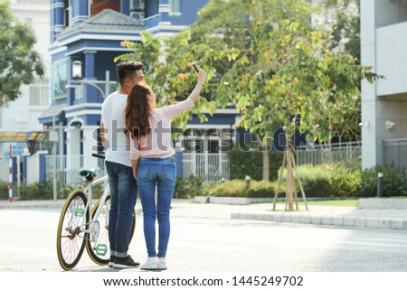Rear view of Asian young couple standing with bicycle on the street and making selfie portrait on mobile phone Royalty-Free Stock Photo #1445249702