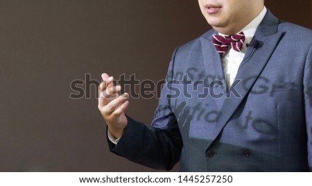 The male speaker with the lavaliere microphone, bow tie and suit is doing the public speaking under the spotlight with grey background.