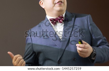 The male speaker with the lavaliere microphone, bow tie and suit is doing the public speaking under the spotlight with grey background.