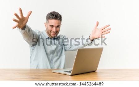 Young handsome man working with his laptop feels confident giving a hug to the camera.
