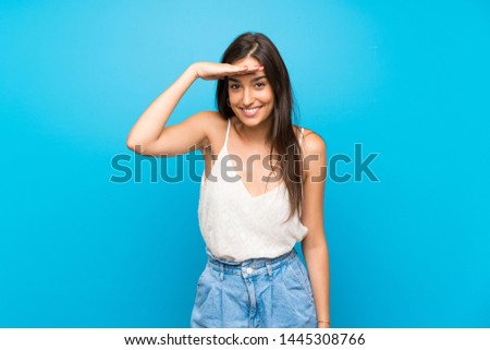 Young woman over isolated blue background looking far away with hand to look something