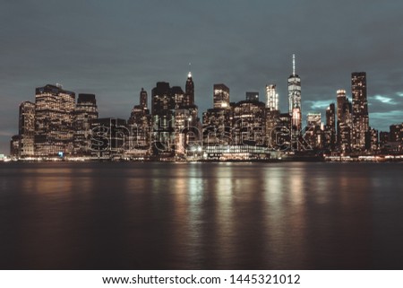 Long Exposure, night photo of Down Town Manhattan from Brooklyn. Hudson River is at the bottom and the buildings with its lights are in the middle of the photo. At top it is a cloudy and grey sky.