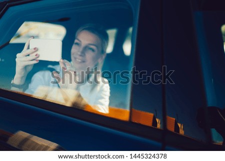 Happy hipster girl sitting at backseat and clicking pictures during car trip using mobile phone camera for photographing, successful Caucasian woman taking images while using automobile service