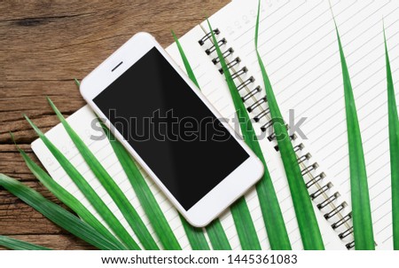White smartphone with black blank mockup screen with green palm leaves and open notebook on the grunge wood desk table . Top view with copy space, mock up flat lay.