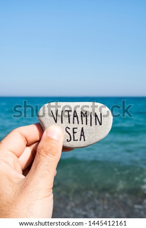 closeup of a caucasian man holding a stone with the text vitamin sea written in it, in front of the ocean