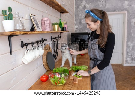 A young woman in an apron with her cat in the kitchen cooks the spring green salad. Summer healthy diet	 Royalty-Free Stock Photo #1445425298
