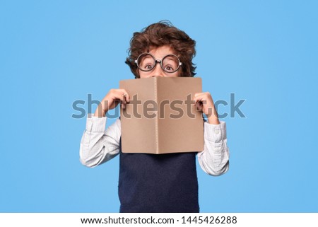 Cute excellent pupil with curly hair and nerdy glasses covering face with open notepad during studies at school against blue background