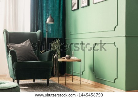 Close-up of a bright, green reading corner of a living room with a gray pillow on a velvet armchair next to a round, wooden coffee table. Real photo