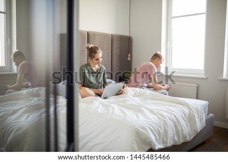 Young spouses using mobile gadgets while sitting on bed in the morning in their bedroom