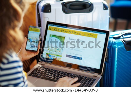 Young tourist woman in striped jacket sitting near couch and trolley bags in the modern house in sunny hot summer day searching for hotel room on online booking website on a laptop and smartphone.