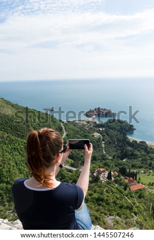 Young white girl making taking a photo with smart phone of Sveti Stefan in Budva, Montenegro. Green cliff and amazing landscape in the adriatic sea