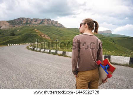 Young stylish man with long hair gathered in a ponytail and in sunglasses stands with a longboard in his hands on a country asphalt road in the mountains on the background of epic rocks