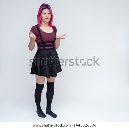 Full-length portrait of young pretty teenager girl in burgundy T-shirt and black dress with beautiful purple hair on a white background in the studio. Talking, smiling, showing hands with emotions.