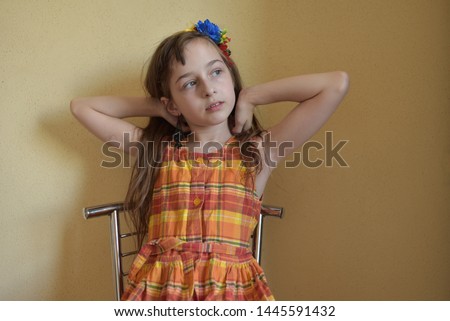Little girl with gray eyes and blond hair sits on a chair. The child sits on a chair with a metal back. A girl of 9 years old in an orange dress. Emotions. Beautiful girl with flowers in her hair.