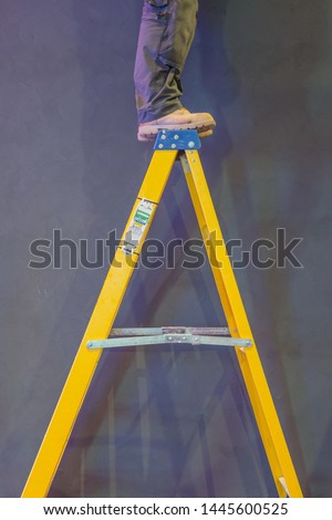 Construction worker standing dangerously on the top rung of a stepladder. Royalty-Free Stock Photo #1445600525