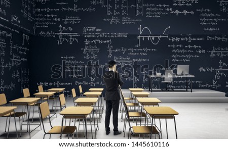 Thoughtful businessman standing in modern classroom interior with furniture and mathematical formulas on wall. Math and complex algorithm concept.