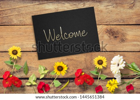 Welcome sign handwritten  on paper envelope on unpainted wooden background in welcoming concept layout with colorful flowers from rustic garden